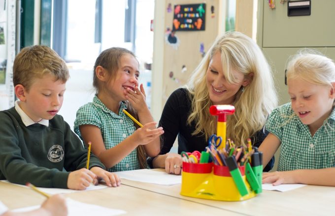 Teacher sits with smiling children at large desk working on paper with pencils in a brightly lit room surrunded by warm colours
