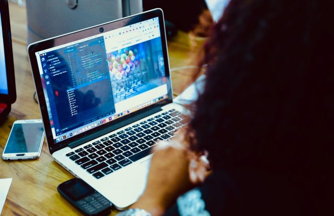 A woman sits at an open laptop with two phones on the desk