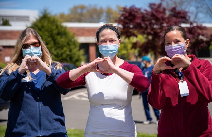 Three nurses standing outside in the sun wearing surgical face masks and holding their hands up in the shape of hearts