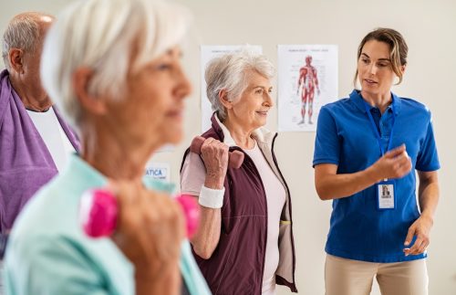Group of old happy people using dumbbells for workout session with physiotherapist. Group of senior man and elderly woman exercising with dumbbells in clinic with trainer. Seniors doing workout weight lifting for wellness with young instructor.