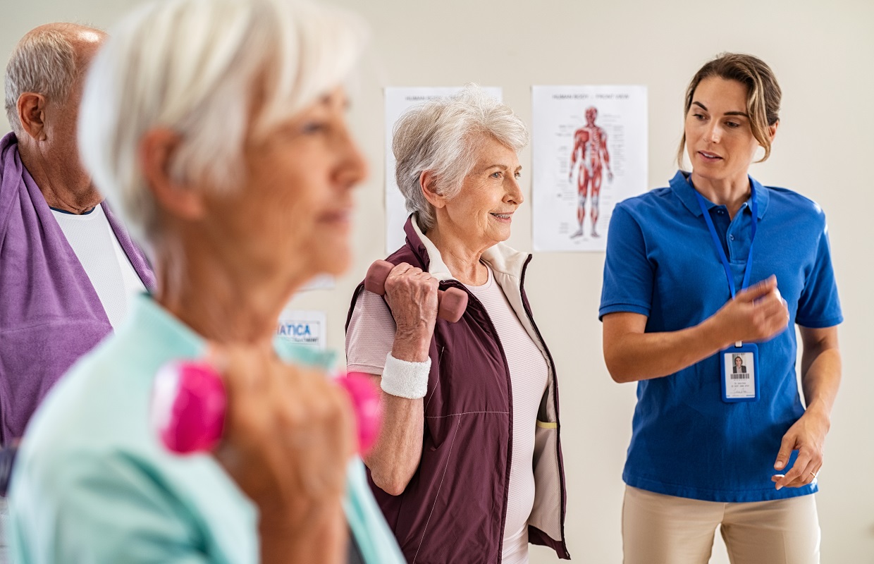 Group of old happy people using dumbbells for workout session with physiotherapist. Group of senior man and elderly woman exercising with dumbbells in clinic with trainer. Seniors doing workout weight lifting for wellness with young instructor.