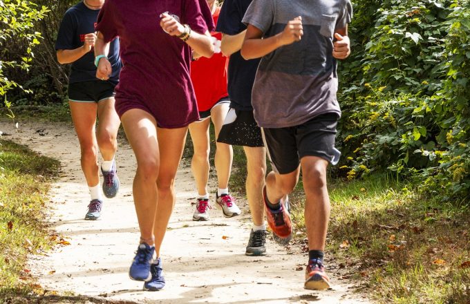 A group of high school runners are training together on a dirt path on a sunny afternoon, running toward the camera.