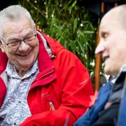 An older man in a red coat laughs on a bench talking to a friend
