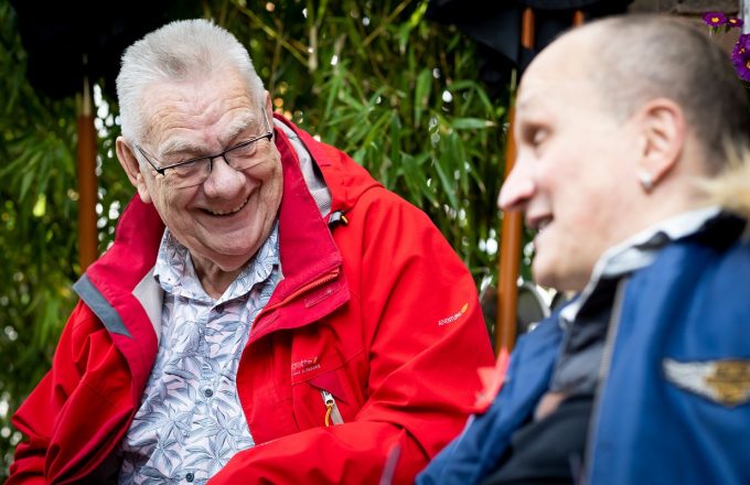 An older man in a red coat laughs on a bench talking to a friend