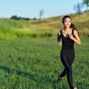 Asian woman in black sportswear running. Japanese runs past camera with flying hair and casts shadow over green grass. Active sport concept