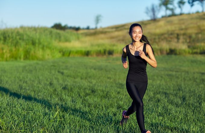 Asian woman in black sportswear running. Japanese runs past camera with flying hair and casts shadow over green grass. Active sport concept