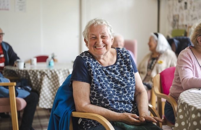 A smiling older woman with a short hairstyle smiles at the camera surrounded by other care homes residents in a living space
