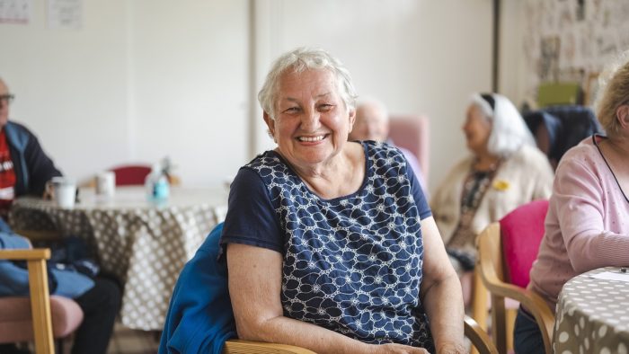 A smiling older woman with a short hairstyle smiles at the camera surrounded by other care homes residents in a living space