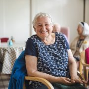 An older lady with short white hair smiles broadly at the camera, seated at a table in a care home settingsmiles broadly at the camera.