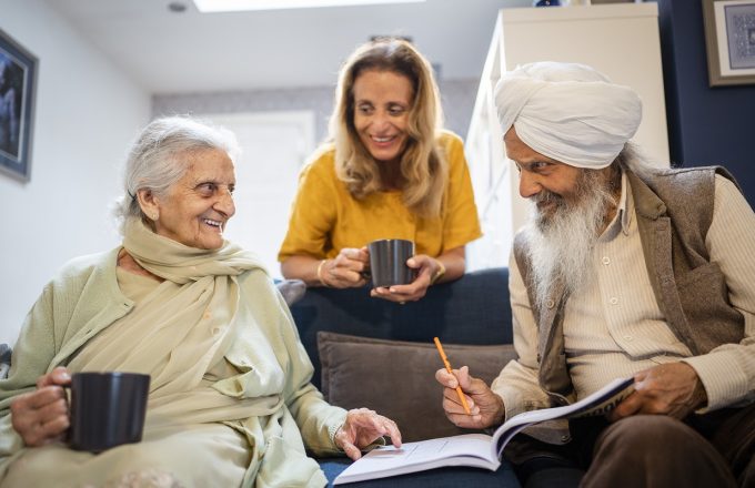 An older South Asian couple sit together on a sofa talking over a cup of tea and a puzzle book. A younger person holding a cup leans on the back of the sofa smiling at them.