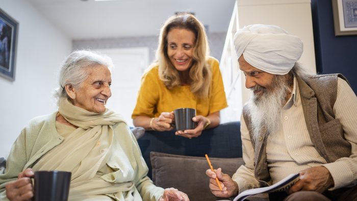 An older South Asian couple sit together on a sofa talking over a cup of tea and a puzzle book. A younger person holding a cup leans on the back of the sofa smiling at them.