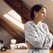 A young woman looks off into the distance, holding her arms crossed against her torso and looking troubled in a domestic space.