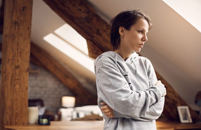 A young woman looks off into the distance, holding her arms crossed against her torso and looking troubled in a domestic space.