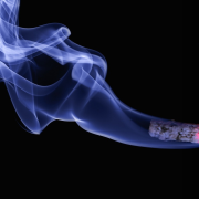 A lit cigarette over a black background with blue-white smoke billowing from the end.