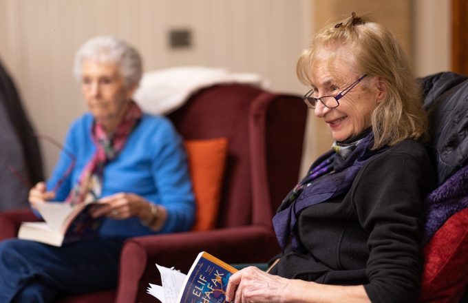 Two older women reading books and smiling in a care home setting.