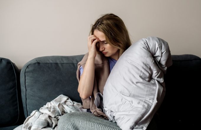 A young woman with long covid syndrome sits on a sofa, clutching her head.