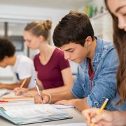 Group of college, secondary school or A-level students studying in classroom writing notes during lesson. Focused guy and girls studying in college library sitting at desk. Group of multiethnic university students doing research sitting in a row.