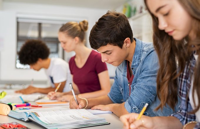 Group of college, secondary school or A-level students studying in classroom writing notes during lesson. Focused guy and girls studying in college library sitting at desk. Group of multiethnic university students doing research sitting in a row.