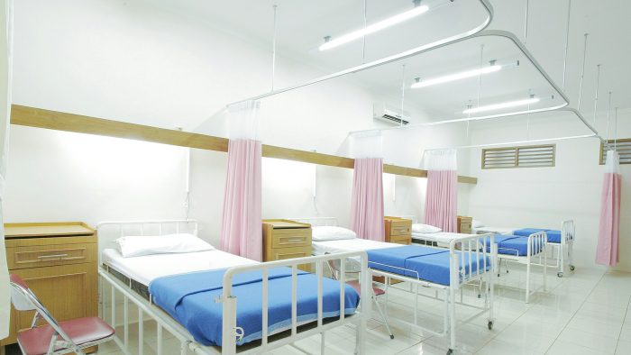 A row of three hospital beds with blue blankets and pink curtains separating each bed.