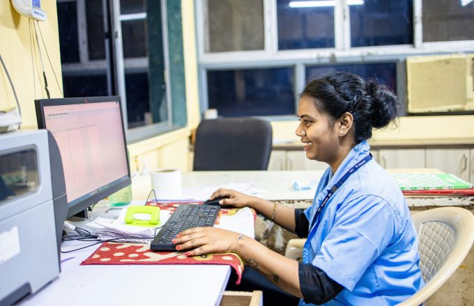 A woman in a blue uniform and a lanyard sits at a desk with a computer, keyboard and mouse