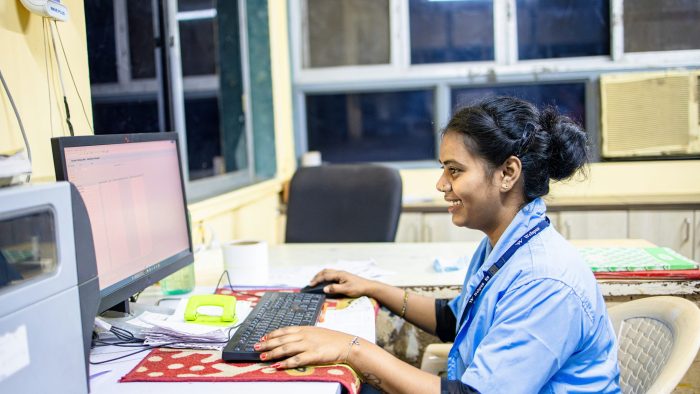 A woman in a blue uniform and a lanyard sits at a desk with a computer, keyboard and mouse
